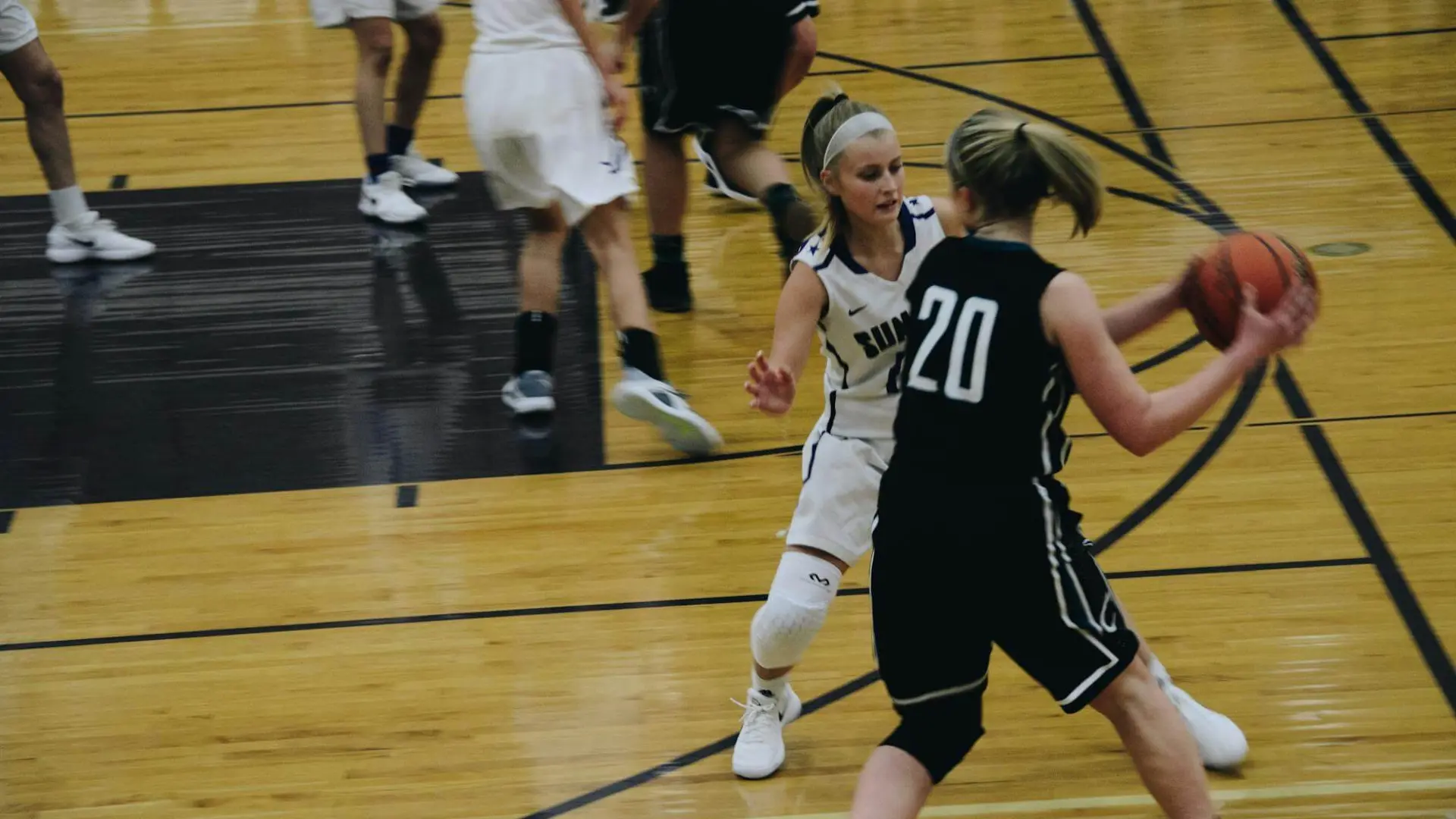 Basketball game, player in black jersey holds ball, player in white defends on a wooden court.