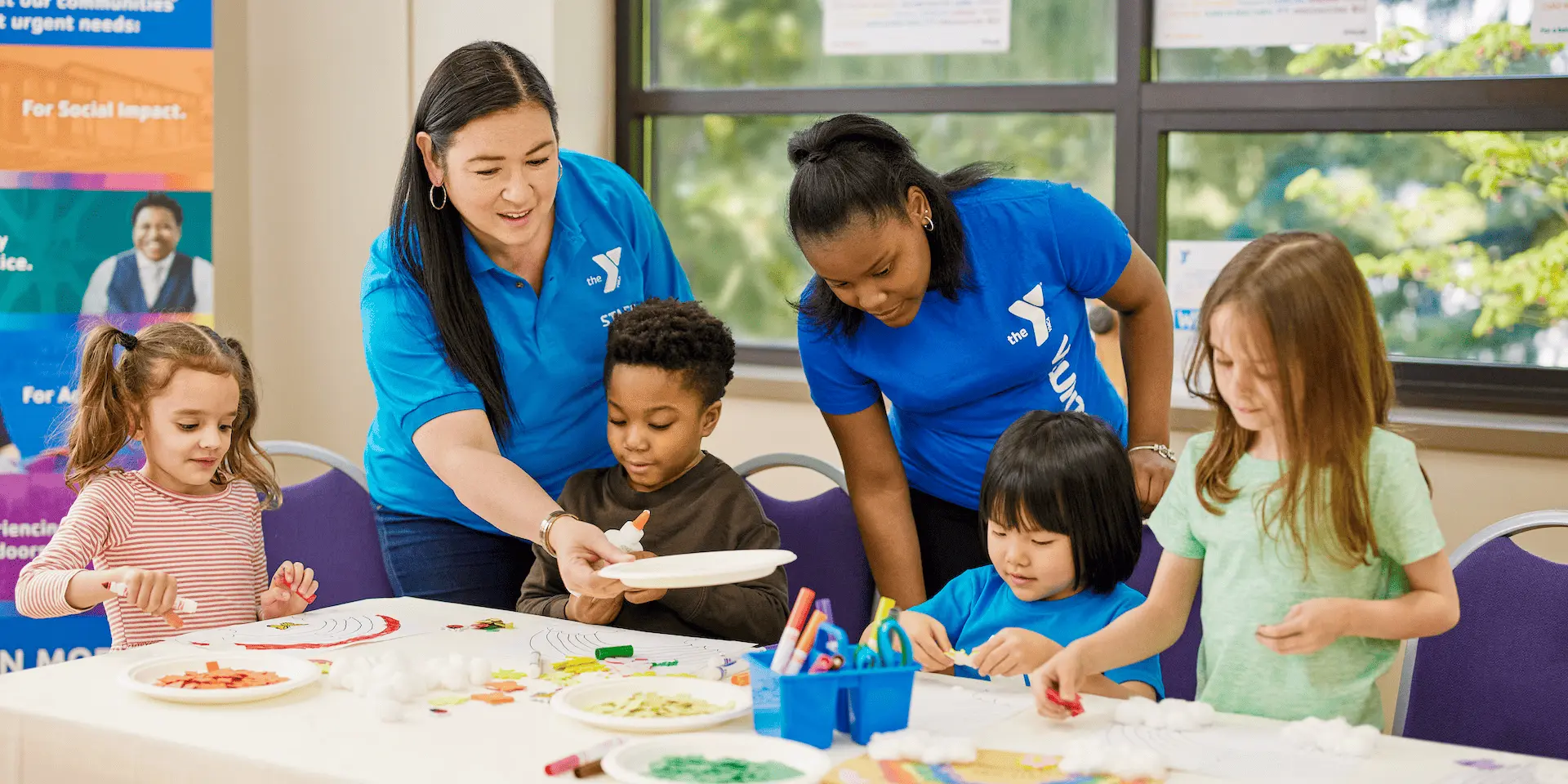 Children and adults crafting at a table, bright room with large windows.