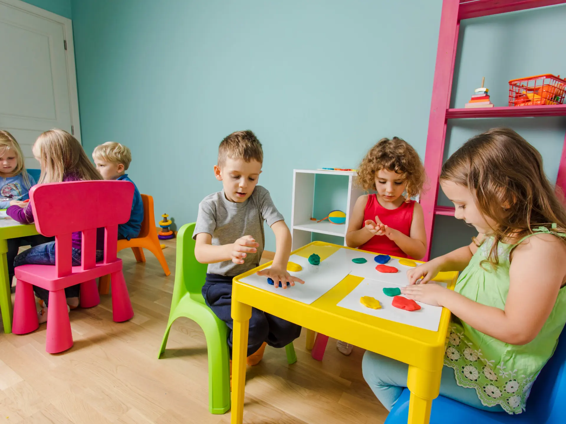 Children crafting at small tables in a classroom, colorful chairs around them.