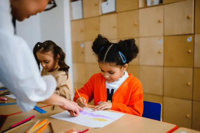 Children drawing with colored pencils at a table in a classroom.