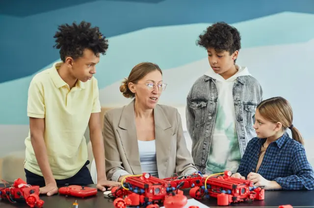 Teacher and three kids discussing red toy models at a table.