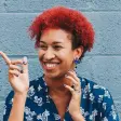Smiling person with curly red hair, wearing a patterned blue shirt, against a brick wall.