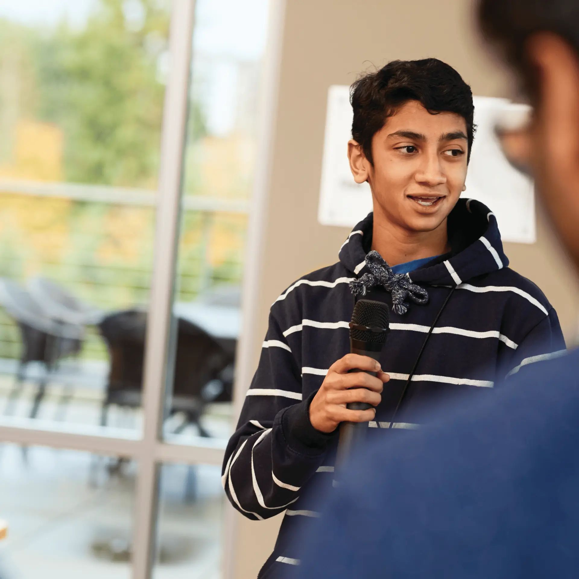 Teen holding a microphone, speaking in a room with blurred people listening.