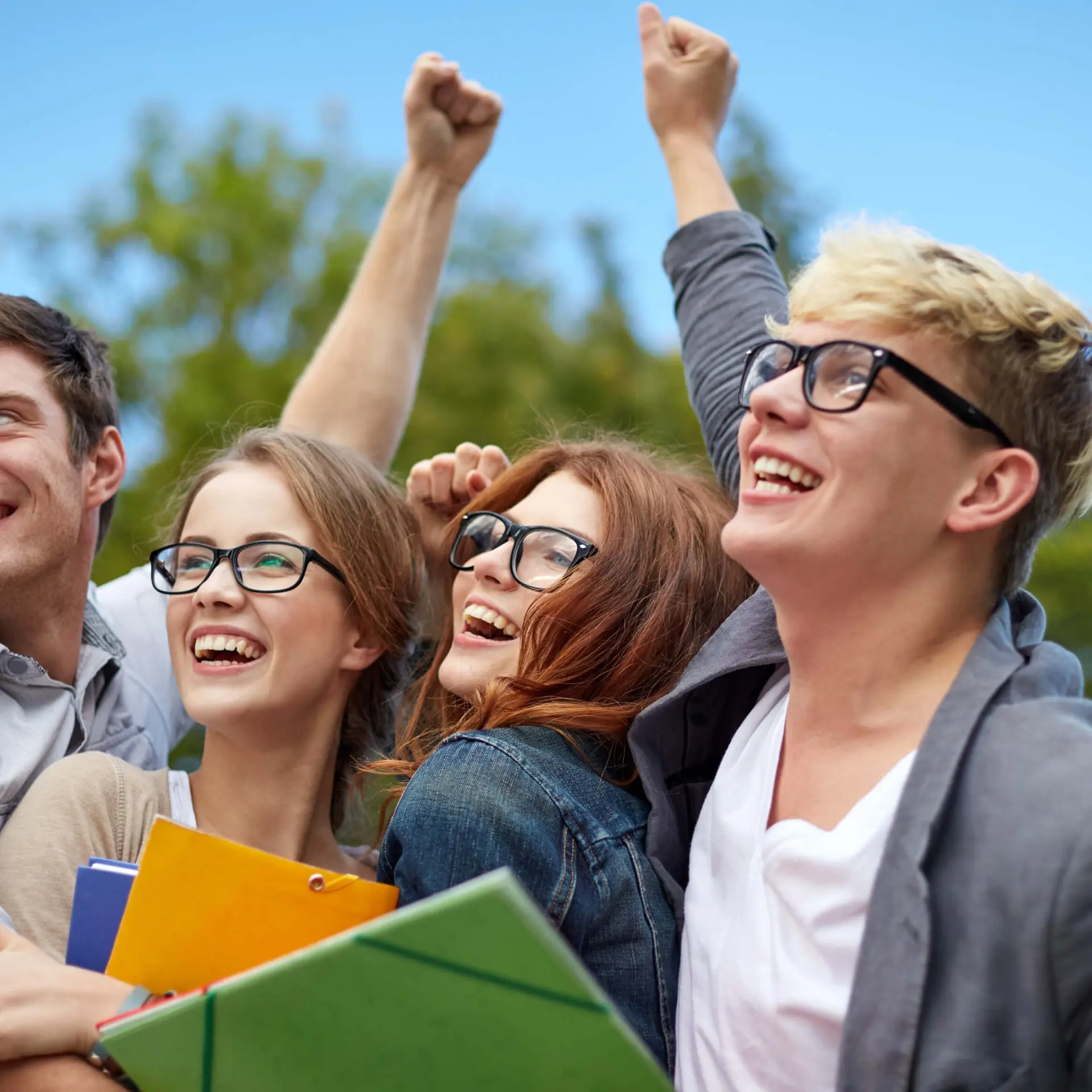 Four smiling friends outdoors, holding books and cheering.